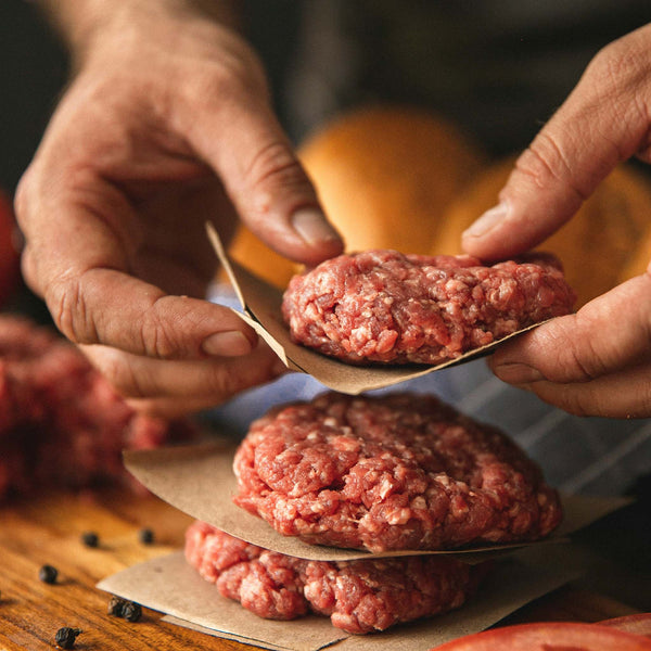 A person preparing raw burger patties, separated by butcher paper, on a wooden cutting board with whole peppercorns.