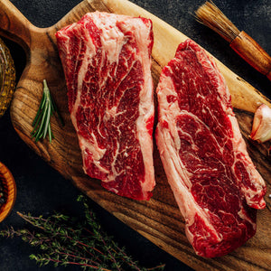 Two raw steaks resting on a cutting board with garlic cloves and fresh herbs.