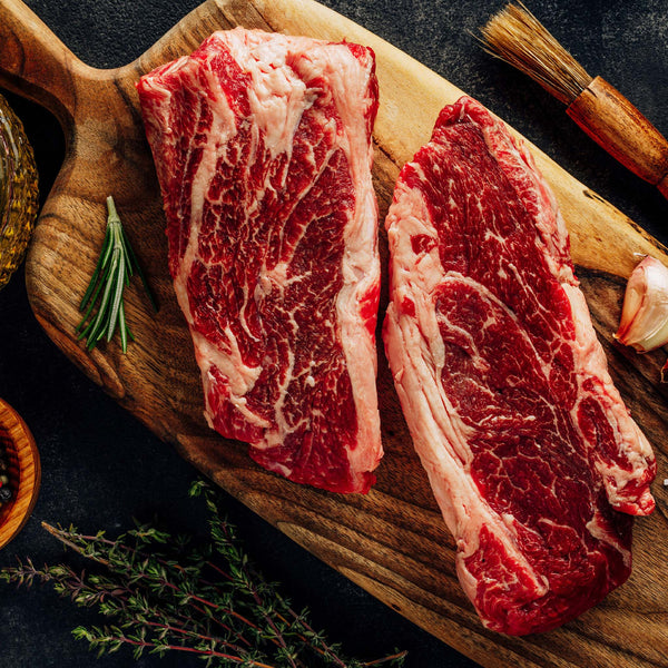 Two raw steaks resting on a cutting board with garlic cloves and fresh herbs.