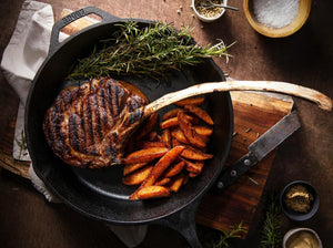 Cooked tomahawk steak in cast iron skillet, with sweet potato fries and fresh herbs, sitting on a wooden cutting board with bowls of seasonings.