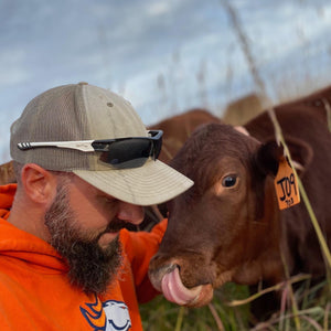 Company owner Josh, wearing a broncos sweatshirt, with his arm around brown cow whose tongue is licking its nose.