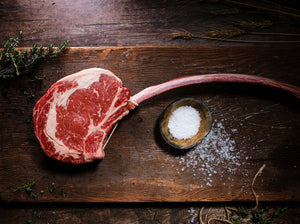 Raw tomahawk steak, resting on a wooden cutting board next to a ramekin of sea salt.
