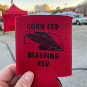 A red Nebraska Cornhuskers Koozie that reads "Corn Fed Bleeding Red" with Memorial Stadium in the background